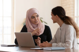 Photo of two people talking and using laptop